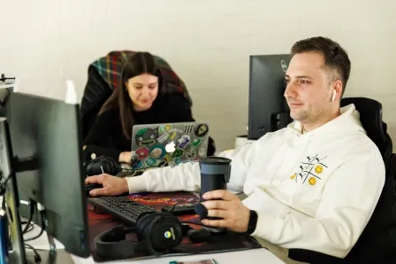 A person in a white hoodie sits at a desk with a computer, wearing earbuds and holding a travel mug.