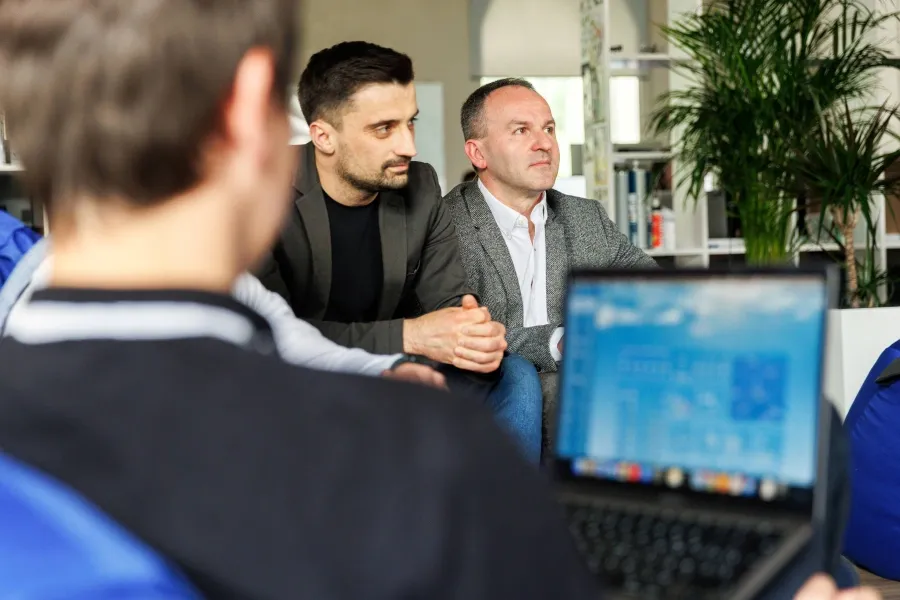 Two men sit attentively in an informal setting, while a person in the foreground uses a laptop.