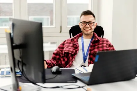 A person wearing glasses and a red checkered shirt sits at a desk with a computer monitor and a laptop.